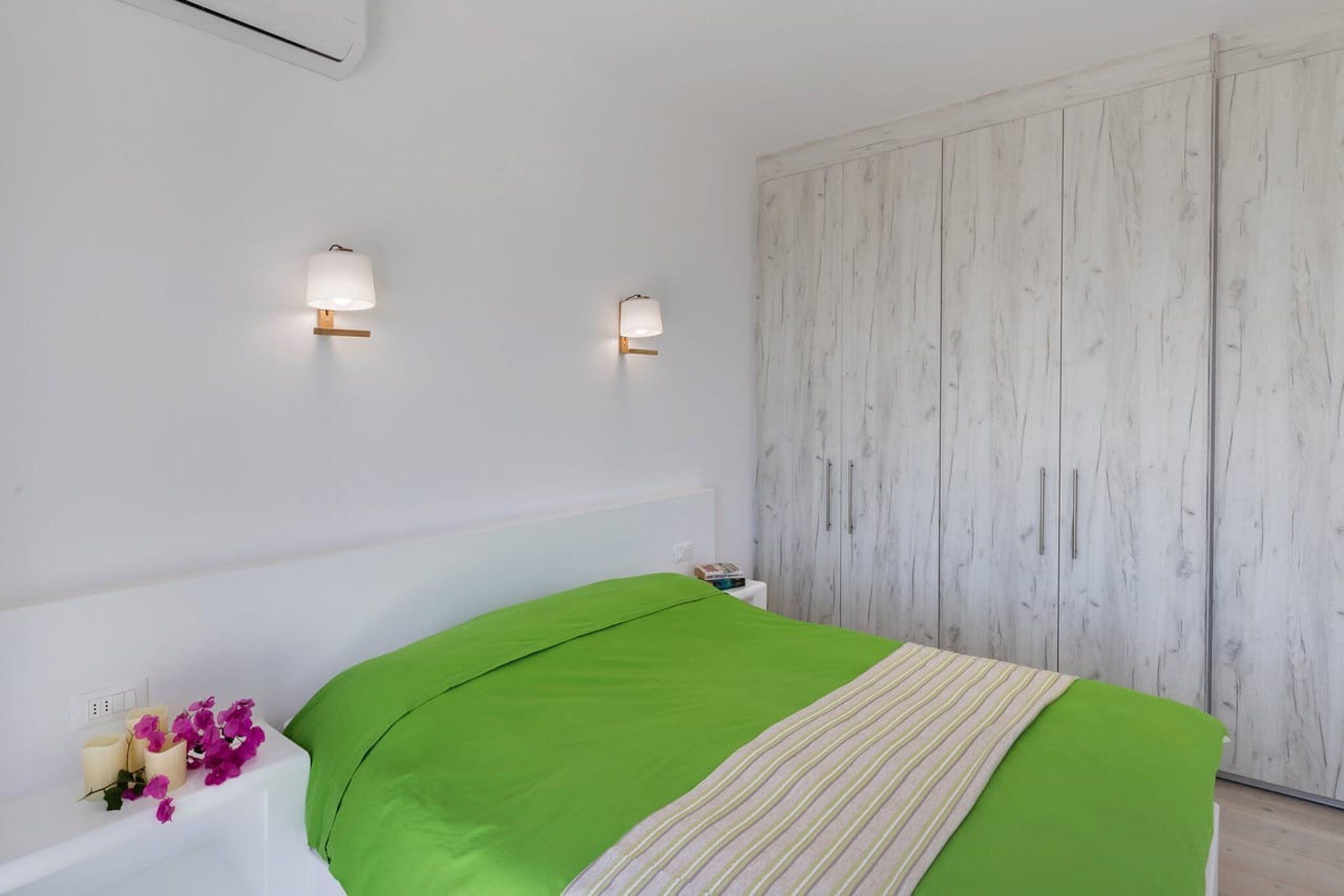 Ein Schlafzimmer mit einem grünen Bettbezug, zwei Wandleuchten, einem weißen Nachttisch und einem Holzschrank.