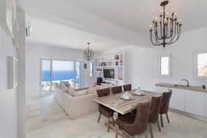 A spacious living area with a large window overlooking the sea, a dining table with chairs, a chandelier, and a white kitchen with a sink and shelves. - Aegle Seafront Villa, 3 Bedrooms, Private Infinity Pool, Agios Stefanos, Syros (Syros)
