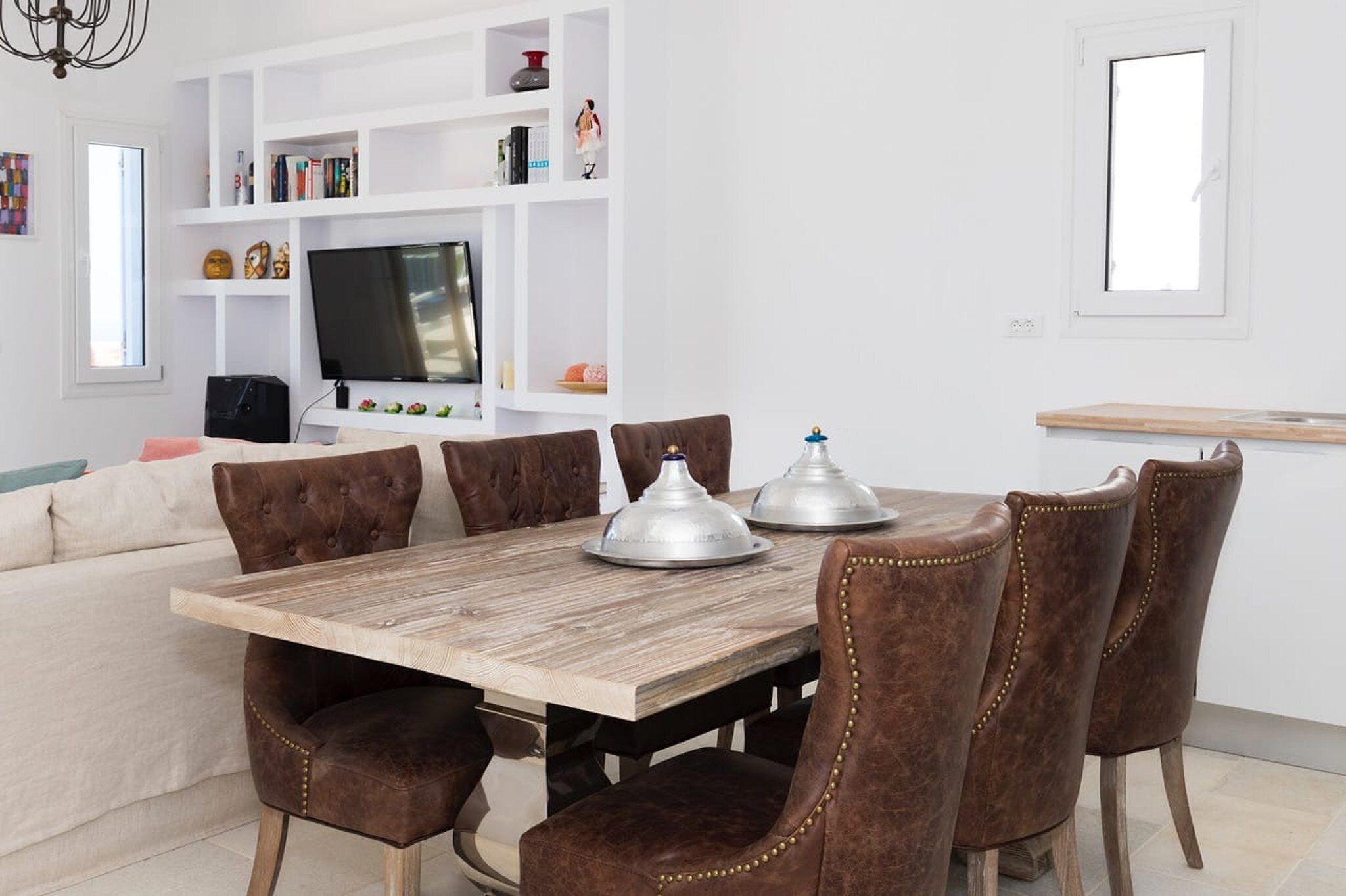 Un comedor con una mesa de madera y sillas tapizadas en tono café, una estantería blanca y un televisor.