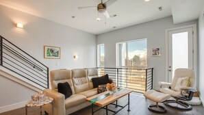 Ein geräumiges Wohnzimmer mit einer Couch, einem Sessel und einem Couchtisch. Eine Treppe mit Geländer, ein Deckenventilator und große Fenster, die natürliches Licht hereinlassen, sind ebenfalls vorhanden.