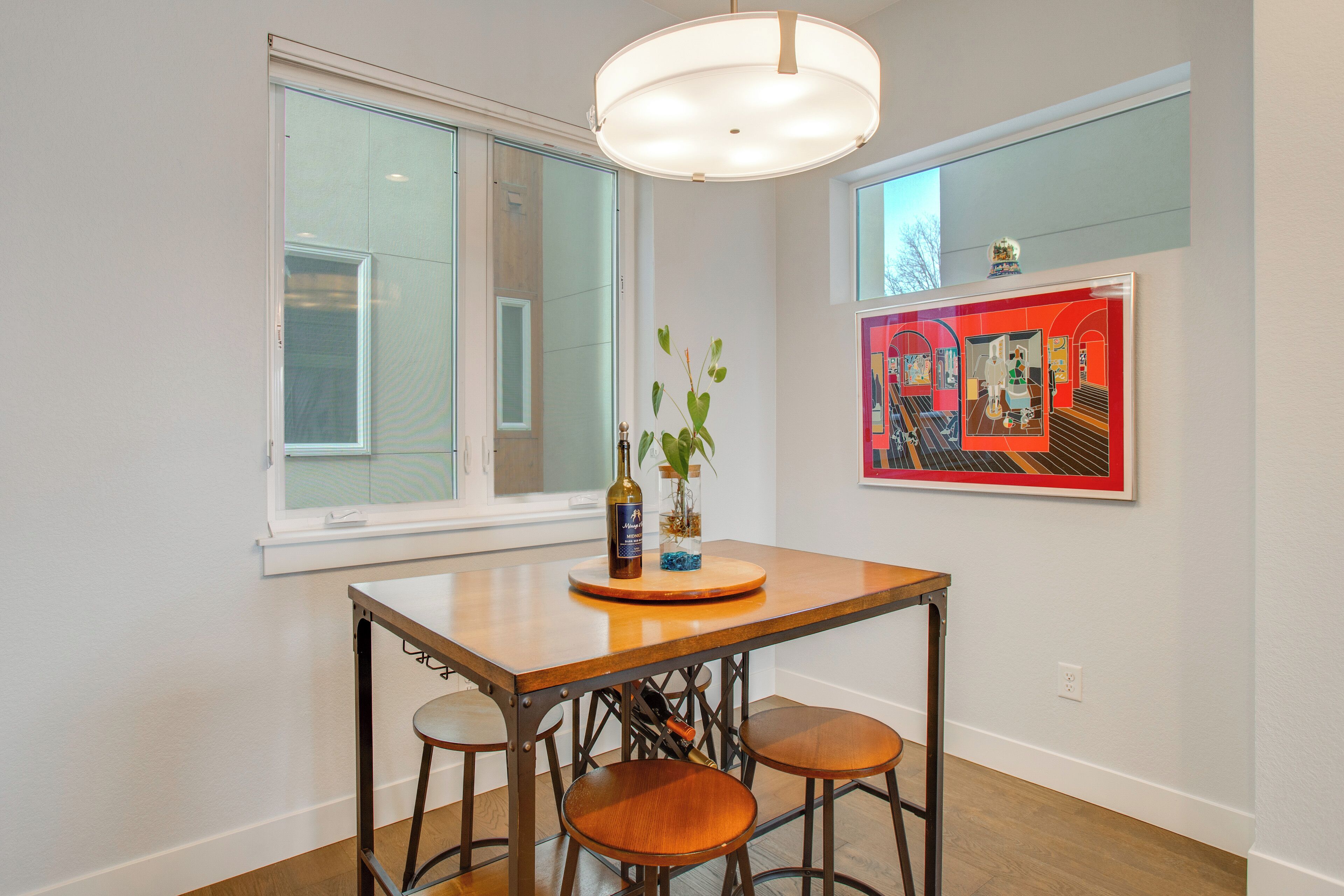 Un coin repas avec une table et des tabourets en bois, un vase avec une plante et une œuvre d’art encadrée accrochée au mur.