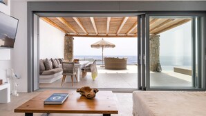 A modern living room with a large sliding glass door leading to an outdoor seating area with a view of the ocean. - Superb Seafront Mykonos Villa | Villa Eros | 4 Bedrooms | Panoramic Sea Views |. (Mykonos)