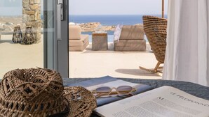 A view from a room with a large window overlooking a beach, featuring wicker furniture and a book. - Superb Seafront Mykonos Villa | Villa Eros | 4 Bedrooms | Panoramic Sea Views |. (Mykonos)