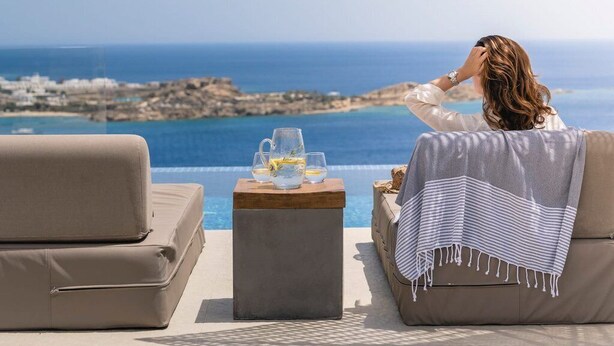 A person relaxing on a beige sofa with a view of the ocean and rocky coastline. - Superb Seafront Mykonos Villa | Villa Eros | 4 Bedrooms | Panoramic Sea Views |. (Mykonos)