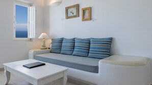 Un salon moderne avec un canapé blanc, des coussins à rayures, une table basse blanche et une petite table d’appoint avec une lampe. Deux tableaux encadrés ornent le mur, et une fenêtre offre une vue sur la mer.