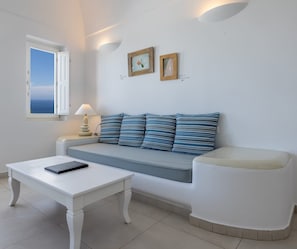 Ein modernes Wohnzimmer mit einem weißen Sofa, gestreiften Kissen, einem weißen Couchtisch und einem kleinen Beistelltisch mit einer Lampe. An der Wand hängen zwei gerahmte Bilder und ein Fenster mit Meerblick.