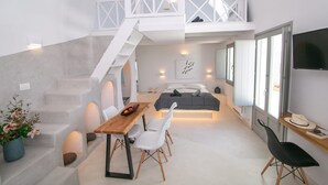 A modern, compact living space with a staircase leading to a loft area, a dining area with a wooden table and chairs, a bed with a gray headboard, and a TV mounted on the wall. - Villa Ioneira, Sea Views, Outdoor Hot Tub, Furnished Terrace, Santorini (Santorini)