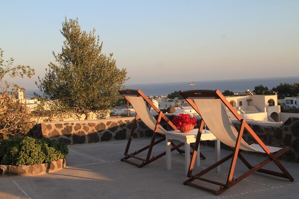 A terrace with two lounge chairs and a table, overlooking a coastal view with buildings and a tree. - Villa Ioneira, Sea Views, Outdoor Hot Tub, Furnished Terrace, Santorini (Santorini)