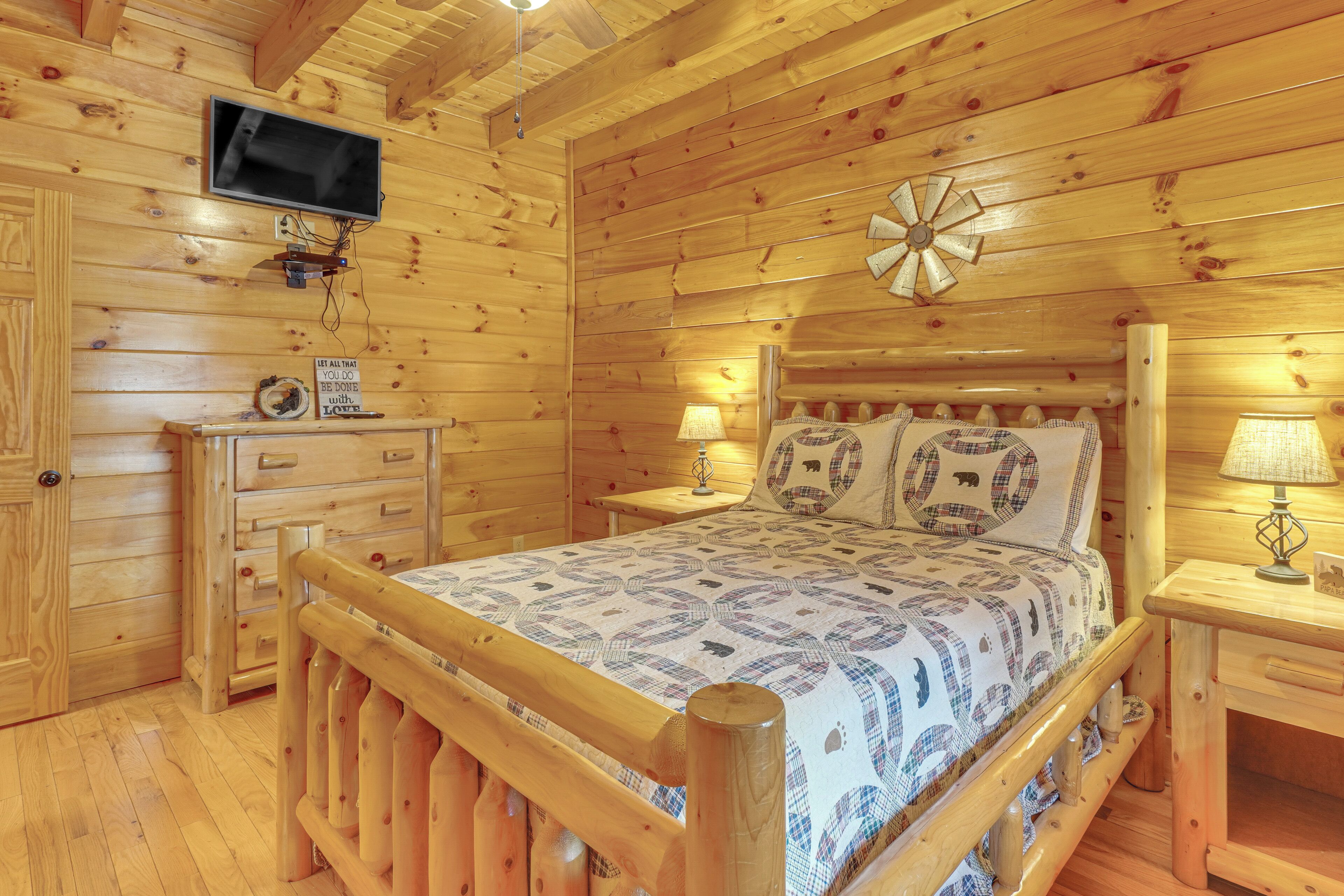 A wooden cabin room with a bed, a television mounted on the wall, and a wooden dresser.