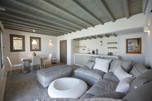A modern living room with a sectional sofa, a dining table with chairs, a white ottoman, and a kitchen area with a countertop and cabinets. - Divine Mykonos Villa Ananke, 4 BDR, Private Pool & Plunge Pool, Sea Views (Mykonos)