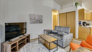 Ein modernes Wohnzimmer mit einem Flachbildfernseher, einem grauen Sofa, einem orangefarbenen Sessel und einem Couchtisch aus Holz.