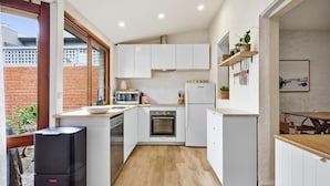 A compact kitchen with white cabinets, a microwave, a stove, and a sink. There is a wooden table and chairs in the dining area, and a small potted plant on the shelf. - The Albert Park Pocket - Stylish Stay with Parking (South Melbourne)