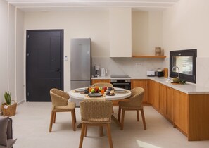 A compact kitchen with a dining area, featuring a round table, wicker chairs, a black door, a stainless steel refrigerator, and a built-in oven. - Villa Evanthia | 2 BDR | Sea Views | Pool | Syros (Syros)