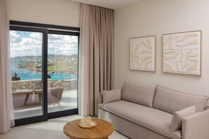 A modern living room with a sofa, a coffee table, and a view of a coastal landscape through large windows. - Villa Evanthia | 2 BDR | Sea Views | Pool | Syros (Syros)