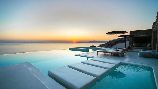 A infinity pool with a view of the sunset over the water, lounge chairs, and umbrellas. - Villa Aqualis, 9 BDR, Sea views, Pool, Elia (Mykonos)