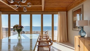 A spacious room with a large glass table, wooden chairs, a vase of flowers, and a view of the sea. - Villa Aqualis, 9 BDR, Sea views, Pool, Elia (Mykonos)
