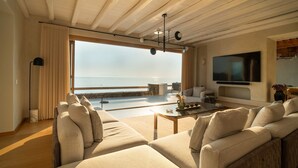 A spacious living area with a large window overlooking the sea, a flat-screen TV mounted on the wall, and a cozy seating arrangement. - Villa Aqualis, 9 BDR, Sea views, Pool, Elia (Mykonos)
