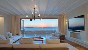 A modern living room with a large TV, comfortable seating, and a view of the sea at sunset. - Villa Aqualis, 9 BDR, Sea views, Pool, Elia (Mykonos)