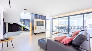 Ein modernes Wohnzimmer mit einer Couch, einem kleinen runden Tisch und einem an der Wand befestigten Fernseher. Eine Treppe führt auf eine obere Ebene, und große Fenster bieten einen Blick ins Freie.