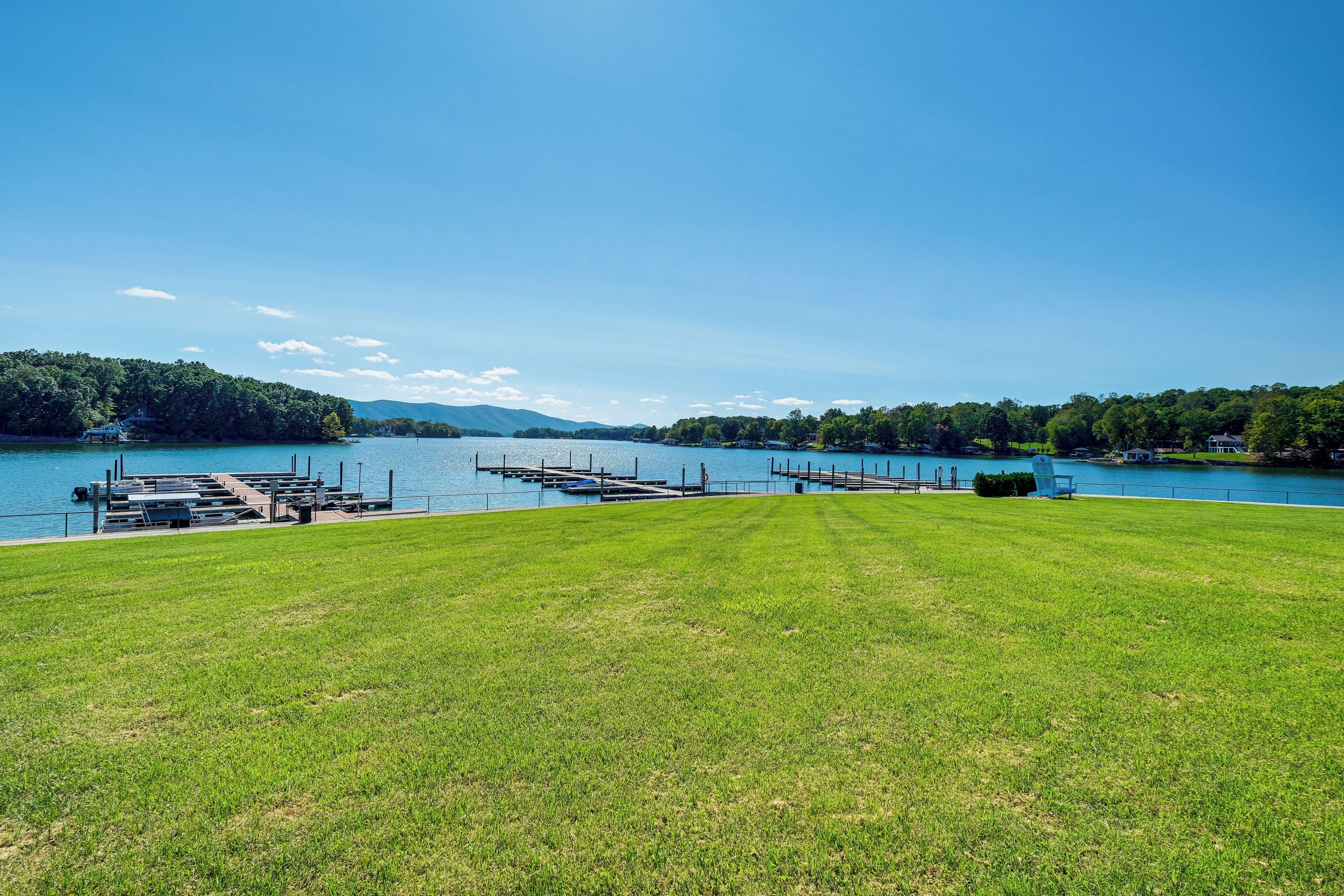Ein grasbewachsenes Feld mit einem Steg, der in einen See führt, umgeben von Bäumen und Hügeln.