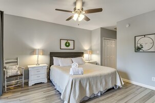 Un dormitorio con cama, dos mesitas de noche, una lámpara de noche, una silla, un ventilador de techo y una pared decorada con un cuadro.