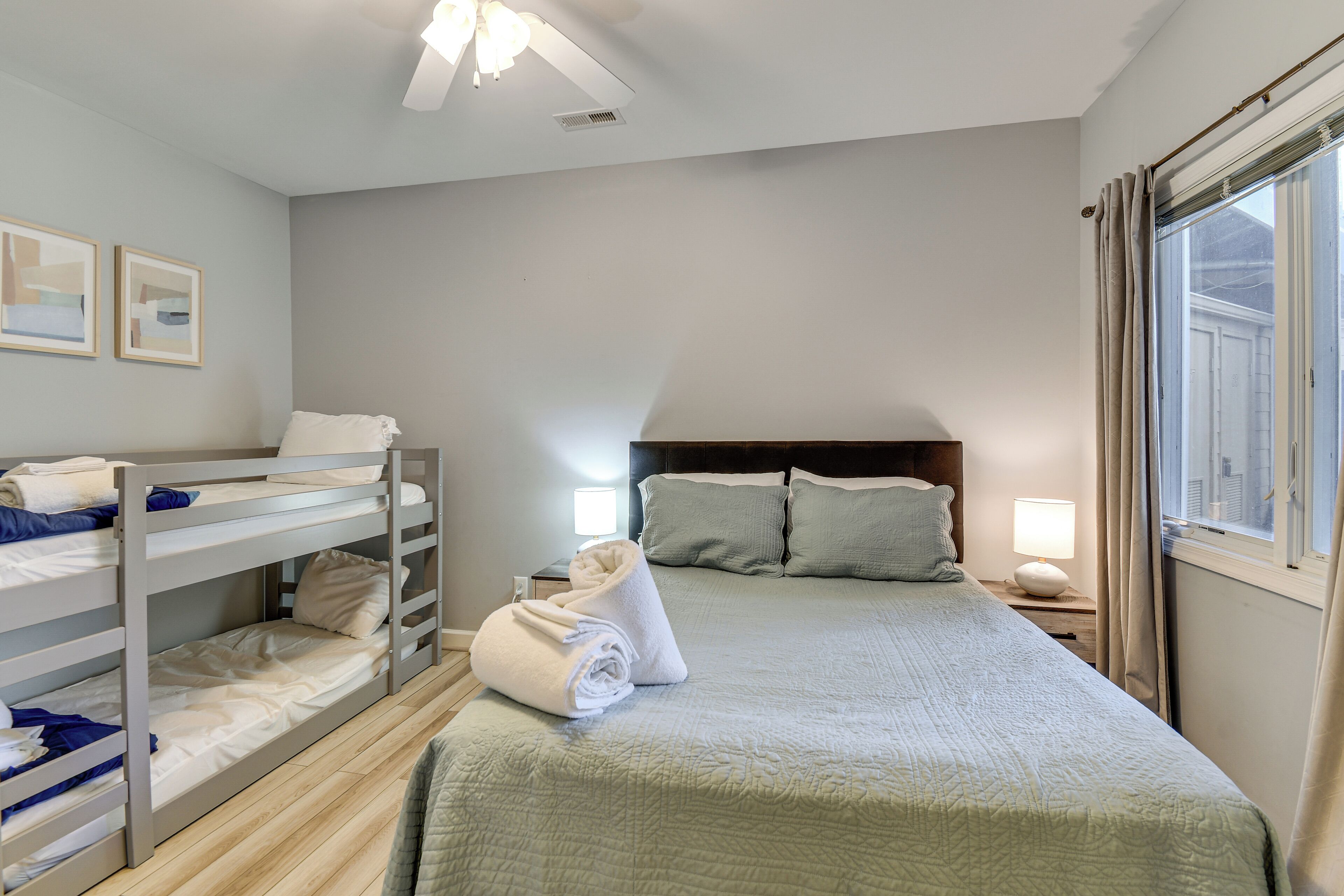 Un dormitorio con literas, una cama grande con colcha, una mesita de noche con lámpara y una ventana con cortinas.