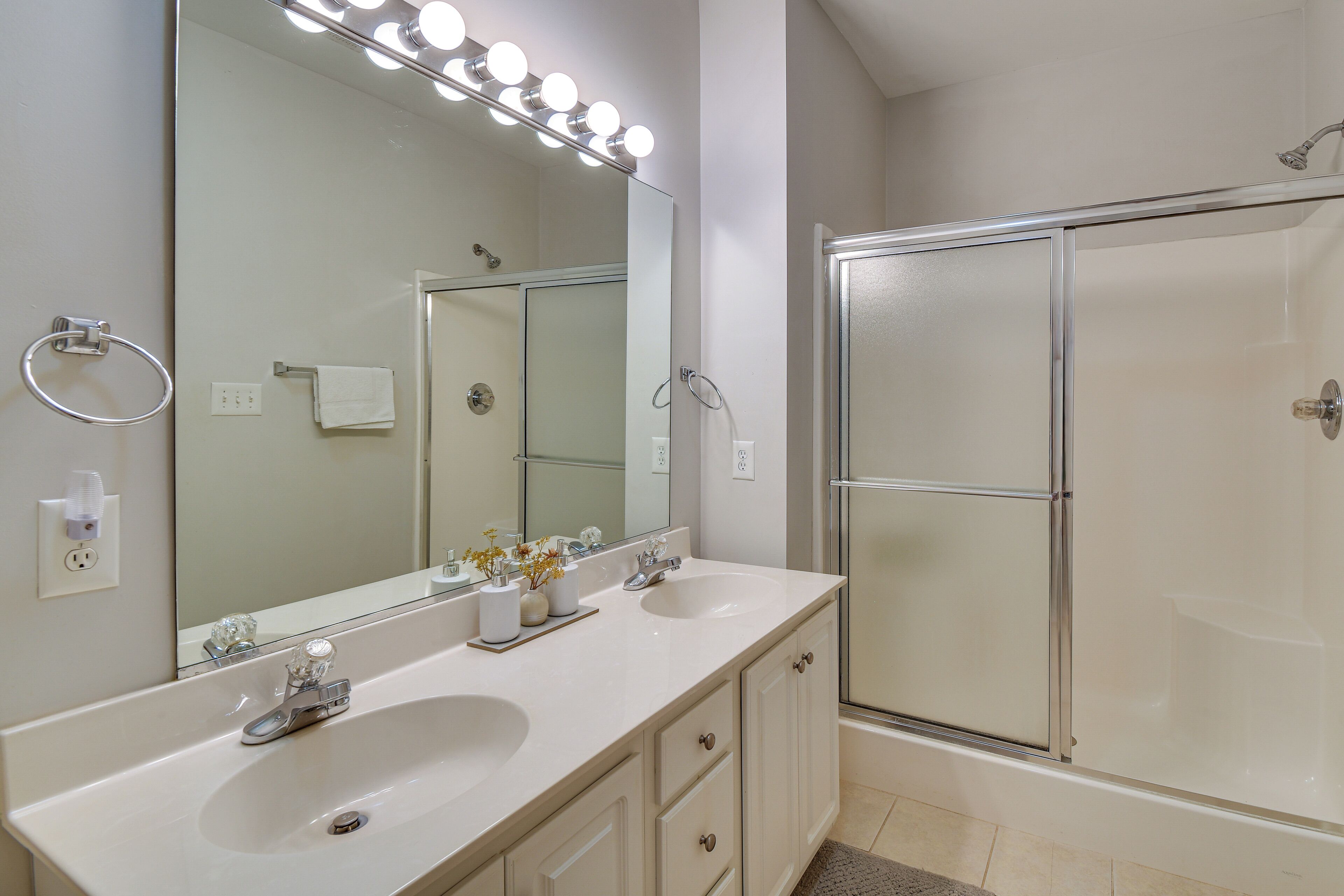 A bathroom with a double sink, a large mirror with lights, a glass shower enclosure, and a towel rack.