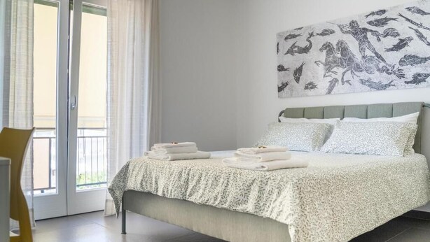 A bedroom with a bed, a chair, a balcony with curtains, and a wall art piece.