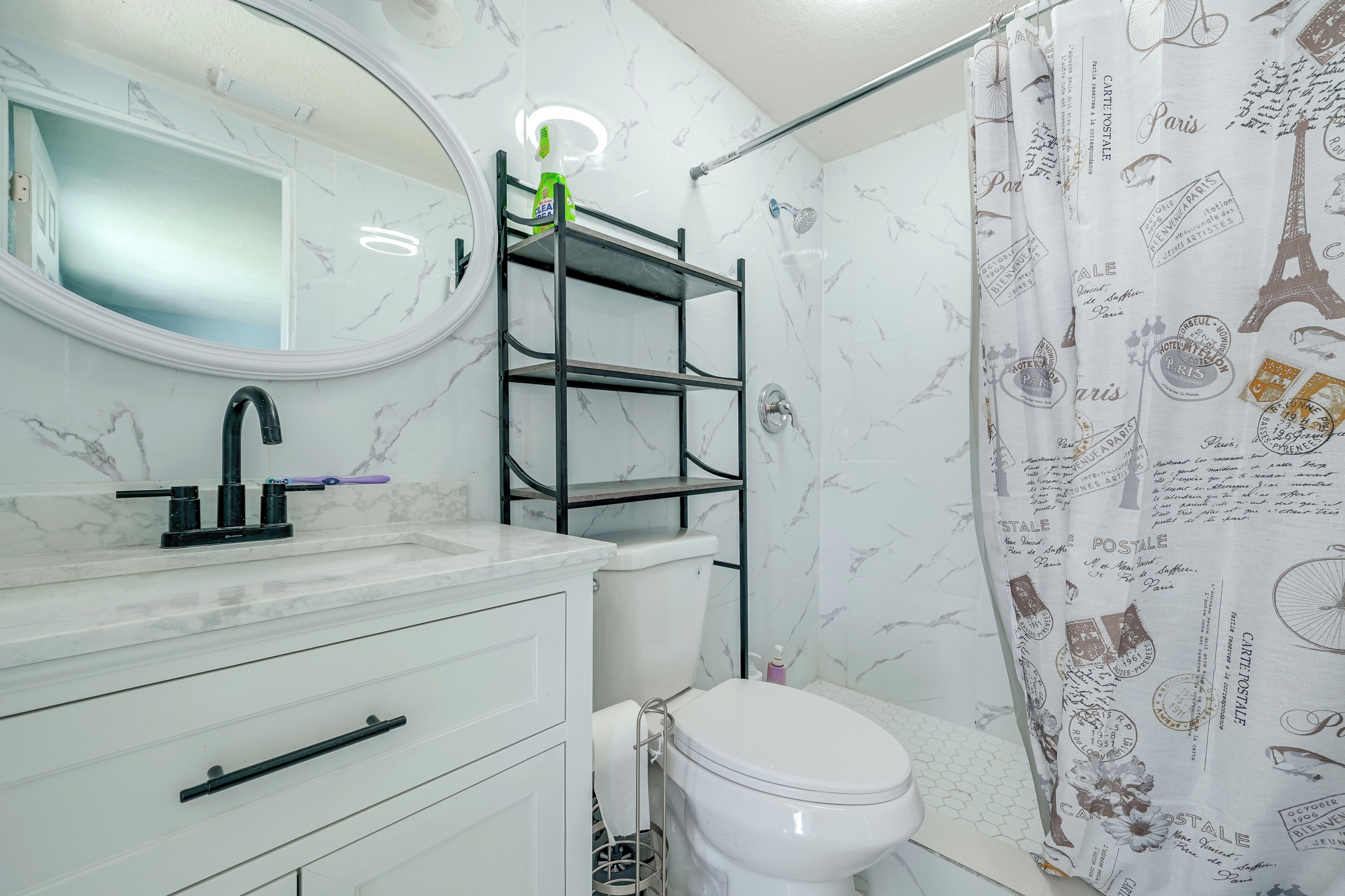 Et badeværelse med en hvid marmorvask, en sort hylde, et hvidt toilet og et bruseforhæng med Paris-motiver.
