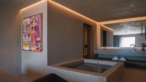 A modern bathroom with a glass-enclosed bathtub, a minimalist sink area, and a colorful abstract painting on the wall. - Rhethis Suite, Divine Sea Views, Infinity Pool, Oia (Santorini)