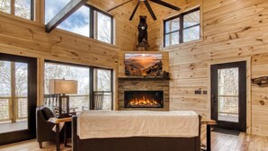 Ein Schlafzimmer in einer Blockhütte mit Kamin, einem Bett, einem Sessel und großen Fenstern.