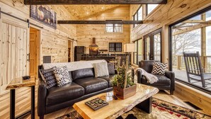 Ein gemütliches Blockhäuschen-Interieur mit Holzvertäfelung, einer schwarzen Ledercouch, einem Couchtisch und einem großen Fenster mit Blick ins Freie.