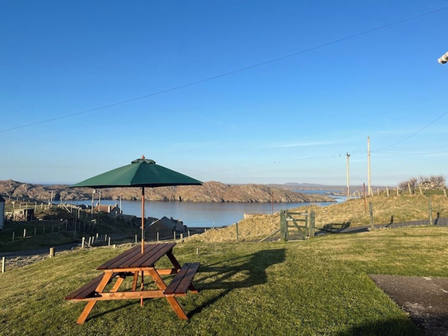 A picnic table with a green umbrella, a grassy area, and a view of a lake with hills in the background.