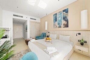 A modern hotel room with a large bed, a bedside table with a lamp, a small seating area, and artwork on the wall. - Seven Palm - 206 Studio (Dubai)