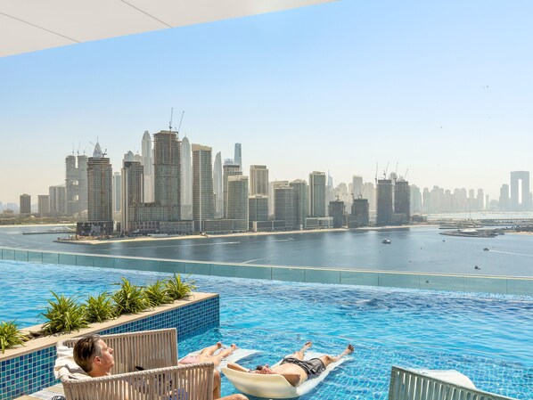 A pool area with a city skyline view, lounge chairs, and a calm water body. - Seven Palm - 206 Studio (Dubai)