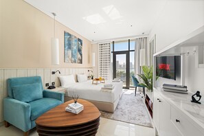 A modern hotel room with a large bed, a blue armchair, a wooden coffee table, a TV, and a city view. - Seven Palm - 206 Studio (Dubai)