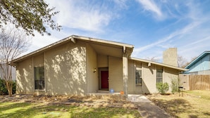 A single-story house with a red door, a covered porch, and a small front yard. - DFW Home Base! Quiet Suburb Stay w/ Yard (Carrollton)