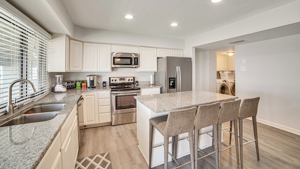 A modern kitchen with white cabinets, stainless steel appliances, a granite countertop, and a bar stool seating area. - DFW Home Base! Quiet Suburb Stay w/ Yard (Carrollton)