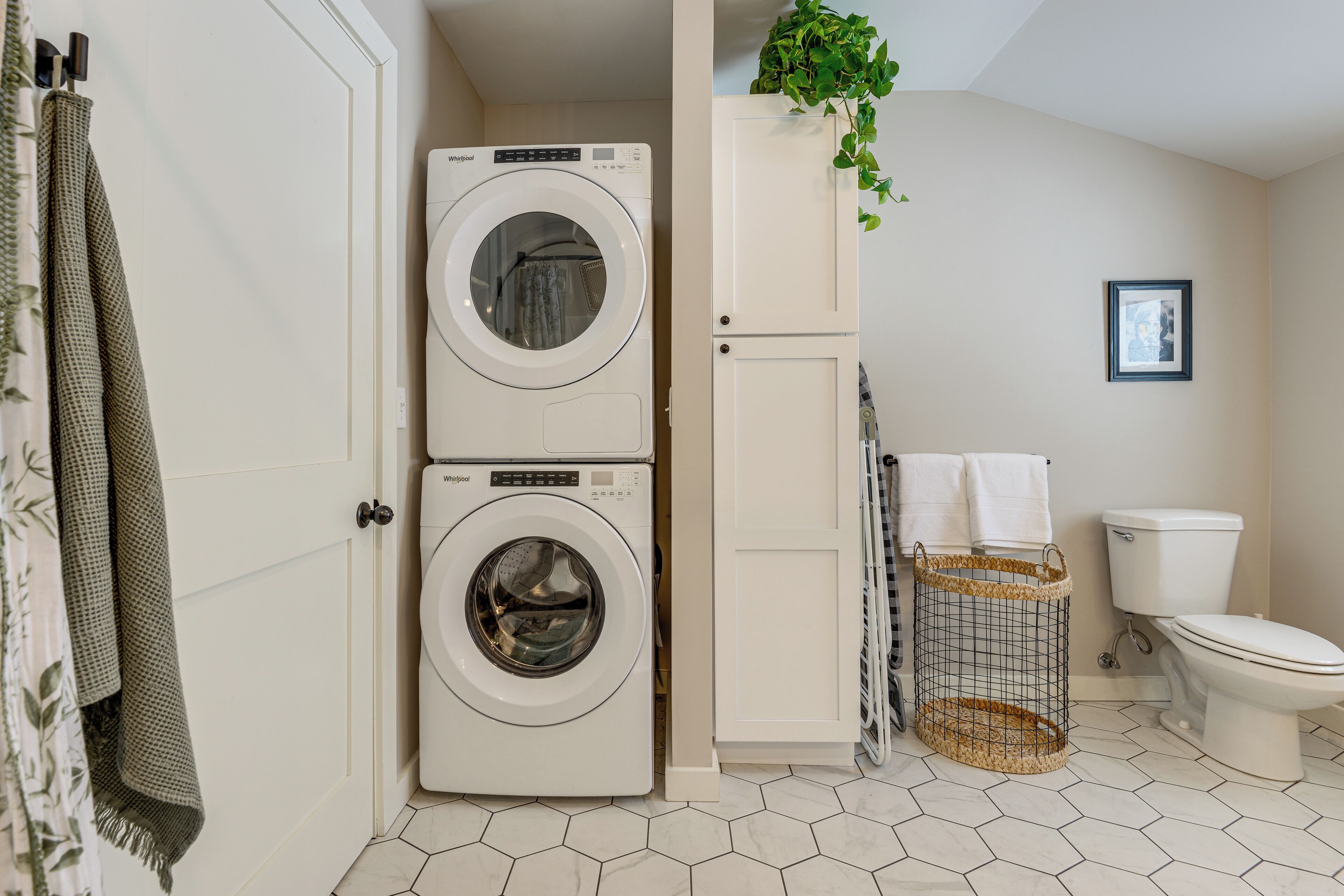 A compact laundry room with a front-loading washer and dryer, a white cabinet, a hanging plant, a framed picture on the wall, and a toilet.
