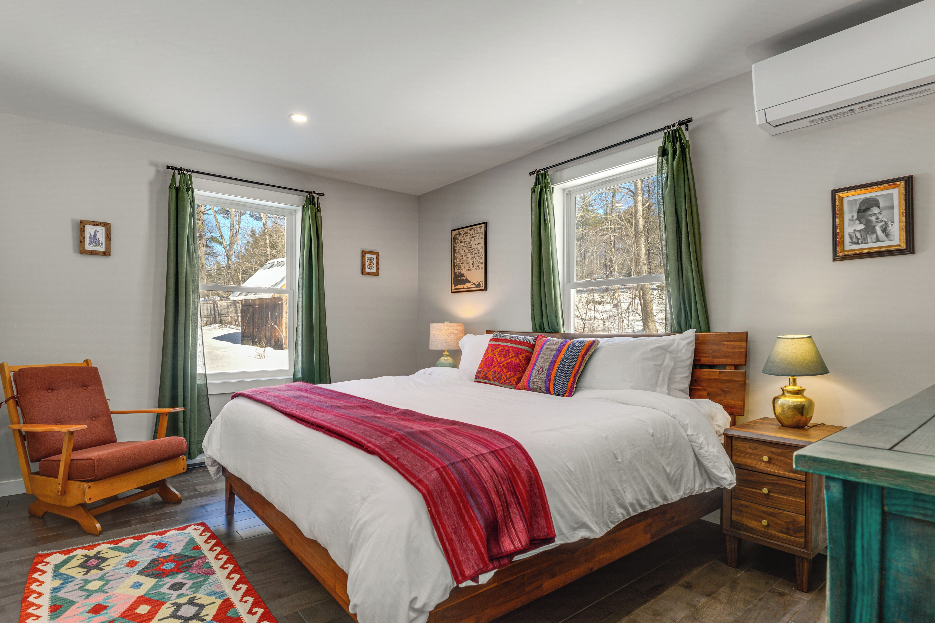 Une chambre à coucher avec un lit en bois, des rideaux verts, un plaid rouge, une table de chevet en bois et une chaise.