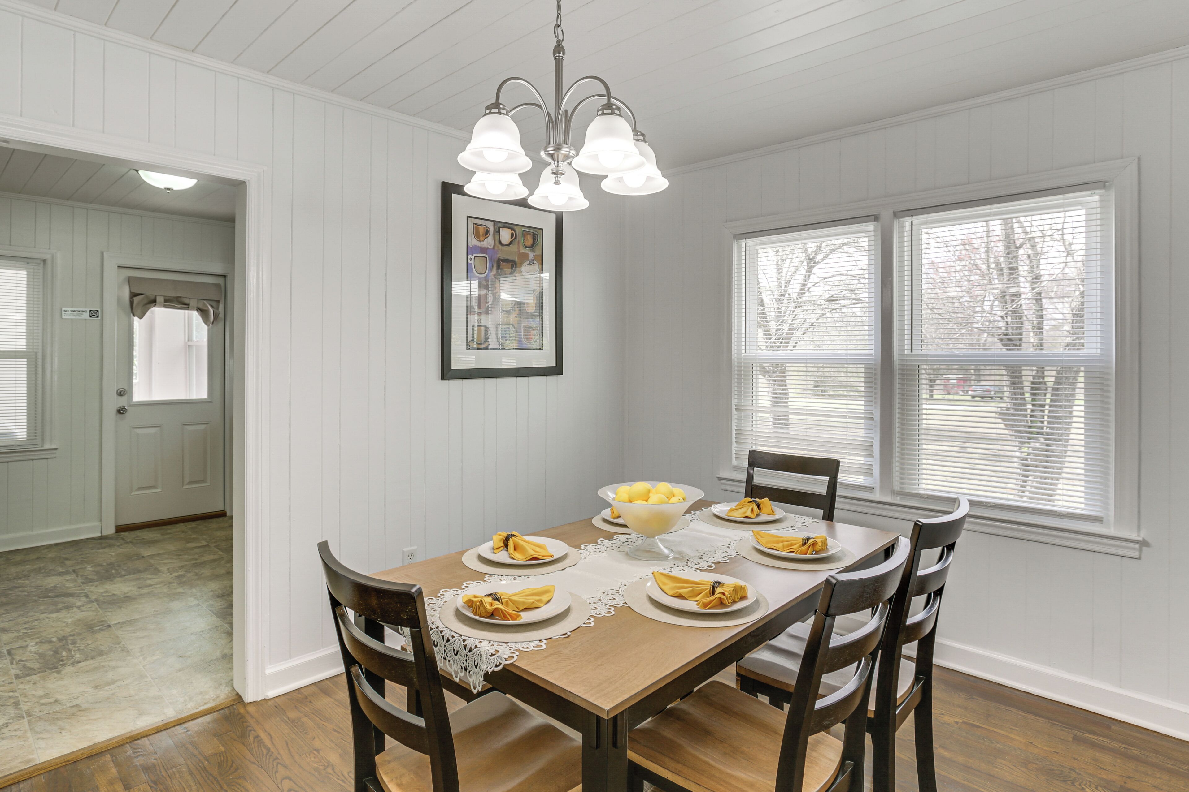 Un coin repas avec une table en bois dressée pour quatre personnes, entourée de chaises. Un lustre est suspendu au-dessus de la table, et une œuvre d’art encadrée est accrochée au mur.