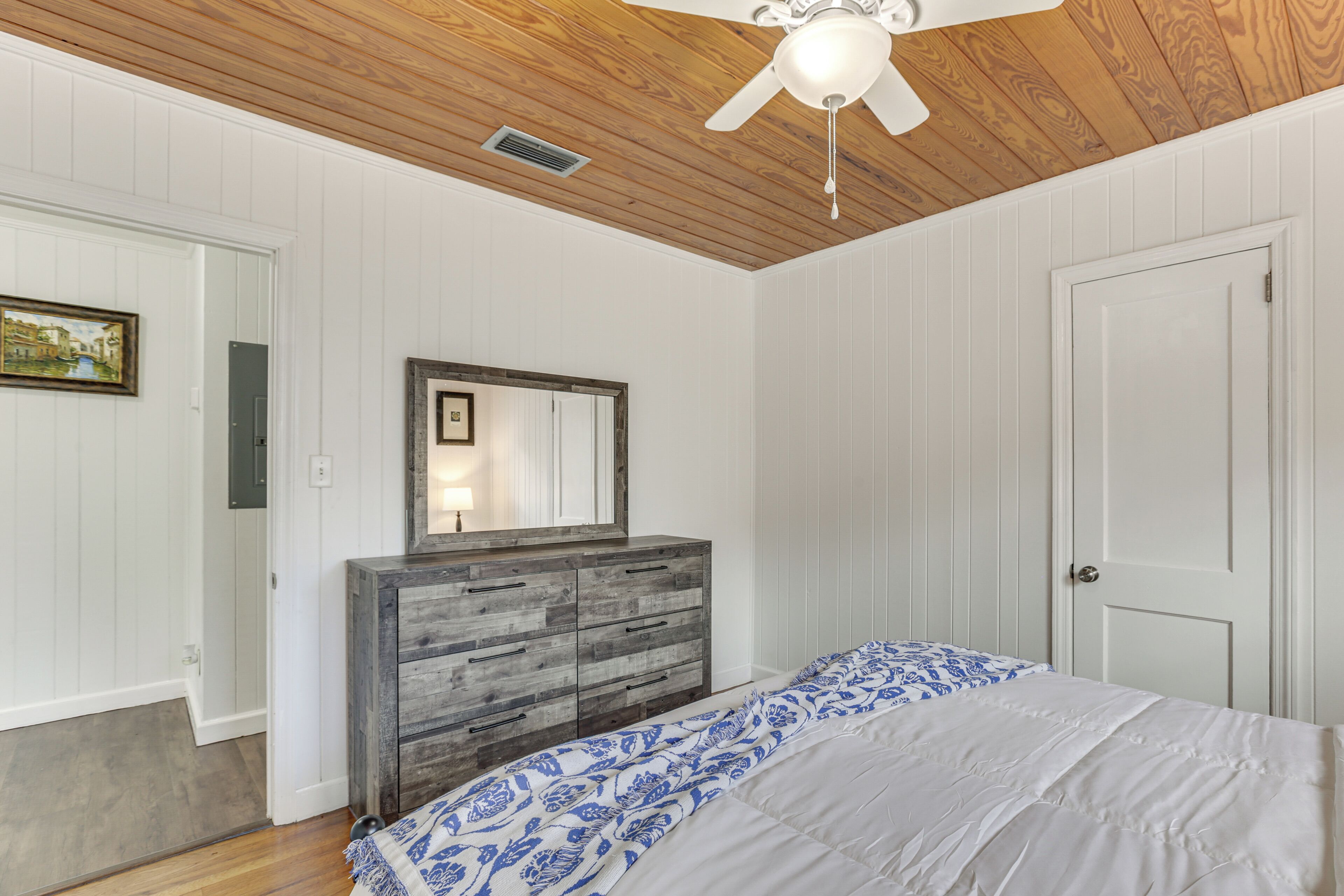 Un dormitorio con techo de madera, una cama con colcha de diseño blanco y azul, un aparador de madera con espejo y una puerta blanca.