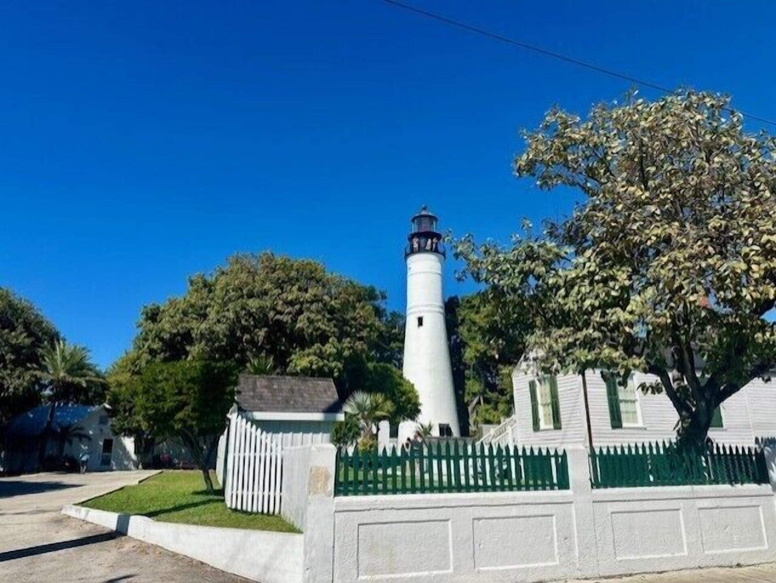 A lighthouse with a white exterior and a black top, surrounded by lush greenery and a white picket fence.