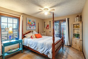 A bedroom with a four-poster bed, a ceiling fan, a window with curtains, a bedside table with a lamp, and a cabinet with decorative items. - Steps to Shore: Group Getaway on Kentucky Lake! (Hardin)