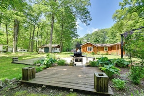 A wooden deck leading to a house with a barbecue area, surrounded by lush greenery and trees. - Rifle Lake Cabin! Michigan Outdoor Adventure Pad (Lupton)