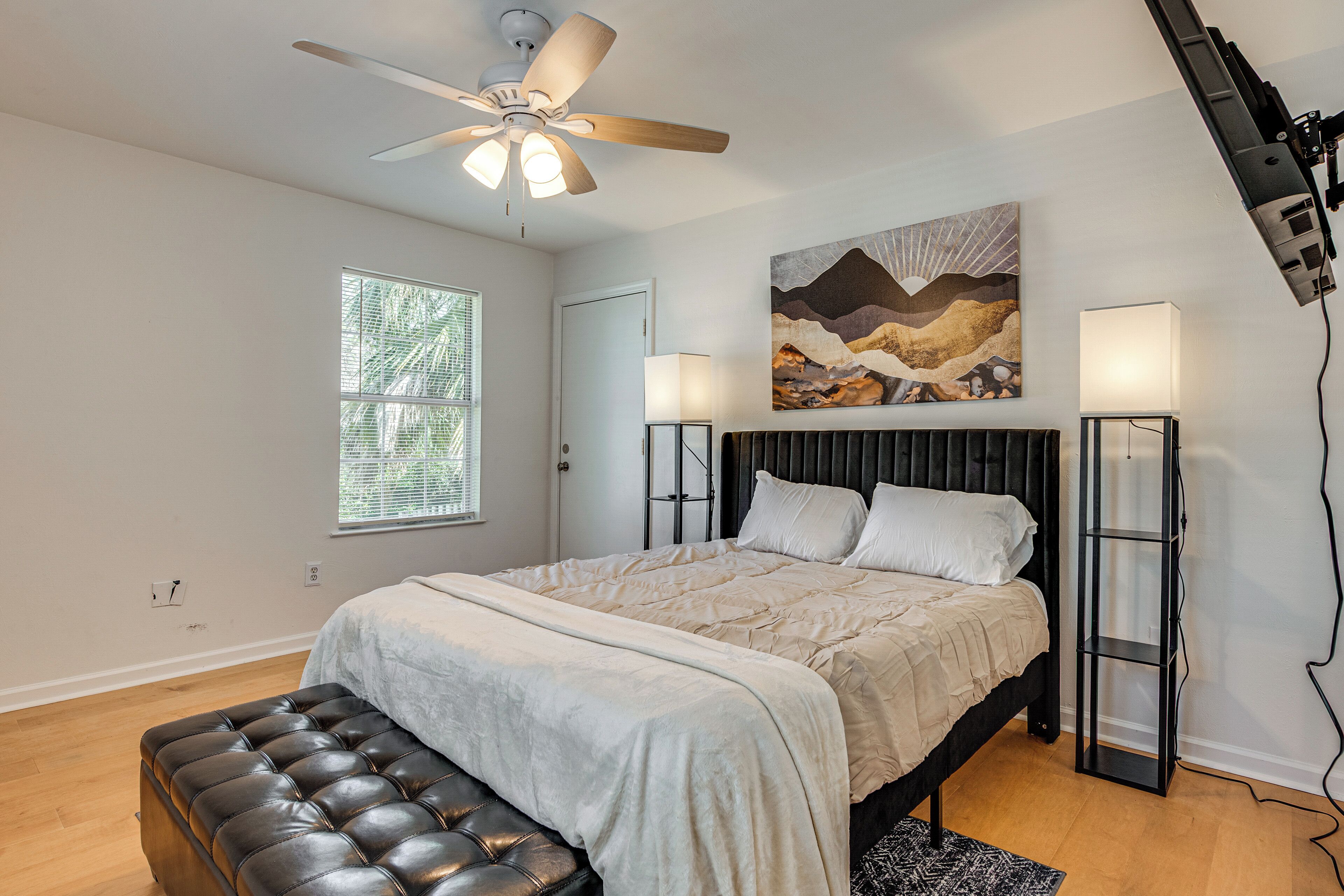 A bedroom with a bed, a ceiling fan, a window, a painting, and a floor lamp.
