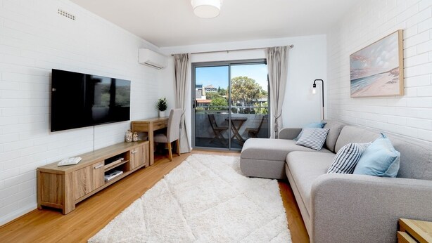 A modern living room with a flat-screen TV, wooden entertainment unit, grey sofa, and a large window with a view of the outdoors. - Stirling Breeze – One Bedroom Apartment Near Beach & Transport (Mosman Park)