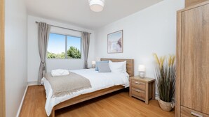 A bedroom with a wooden bed, bedside tables, a wardrobe, a window with curtains, and a potted plant. - Stirling Breeze – One Bedroom Apartment Near Beach & Transport (Mosman Park)