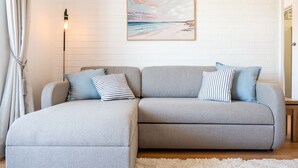 A grey sectional sofa with striped pillows, a white fluffy rug, a floor lamp, and a beach-themed painting on a white brick wall. - Stirling Breeze – One Bedroom Apartment Near Beach & Transport (Mosman Park)