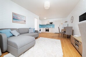 A modern living room with a grey sofa, a white rug, and a kitchen in the background. - Stirling Breeze – One Bedroom Apartment Near Beach & Transport (Mosman Park)
